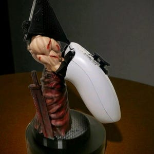 Può includere: Statuetta dettagliata di un personaggio della serie di videogiochi Silent Hill, che tiene in mano un controller di gioco bianco. La statuetta presenta un elmo nero a forma di piramide, un abito rosso e marrone e una grande spada. La base è nera.