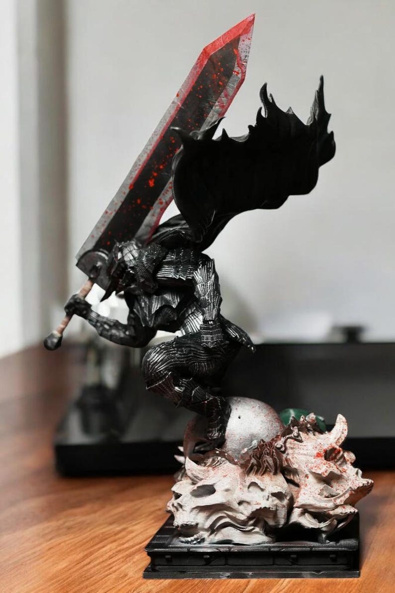 Puede incluir: Una figura de acci&oacute;n negra y plateada de un personaje empu&ntilde;ando una gran espada con salpicaduras de sangre roja. La figura est&aacute; de pie sobre una base hecha de cr&aacute;neos y huesos.