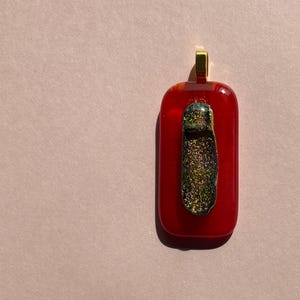 Puede incluir: Un colgante rectangular de vidrio rojo con una fianza dorada. Un elemento vertical de vidrio texturizado, de color verde oscuro y dorado, está centrado en el colgante. El colgante es probablemente para un collar.