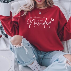 May include: A red sweatshirt with the text "solo una chica que ama Navidad" in white script. The sweatshirt is paired with light blue jeans with distressed knees. A white mug is held in the left hand.