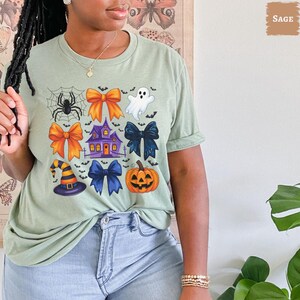 May include: Sage green t-shirt featuring a colorful Halloween-themed graphic. The design includes a spiderweb with a spider, bows, a haunted house, a ghost, a witch's hat, a pumpkin, and bats.
