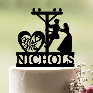 Op de afbeelding: Zwarte silhouet-taarttopper met een stel op een hoogspanningslijn met een hart waarop "Mr. & Mrs. NICHOLS" staat.