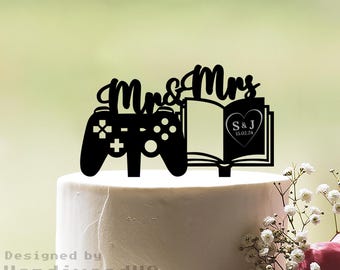 Op de afbeelding: Zwarte taarttopper met de woorden "Mrs & Mrs", een videogamecontroller en een open boek. Het boek toont initialen en een datum in een hart. Geplaatst op een witte taart.