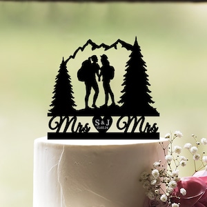 Może przedstawiać: Czarny topper do tortu w kształcie sylwetki dwóch osób wędrujących po górach z sosnami po obu stronach. Topper zawiera napis "Pani S&J 15.02.24 Pani."