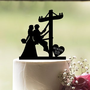 May include: Black silhouette cake topper of a bride and groom. The groom is climbing a utility pole. The bride is holding a bouquet of flowers. A heart with the text "AUSTIN 16.06.2025" is on the right side of the topper.
