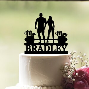 Könnte beinhalten: Schwarzer Silhouetten-Kuchentopper eines Brautpaares, das eine Langhantel hebt. Der Topper trägt die Aufschrift "Mr. & Mrs. Bradley" auf der Langhantel.