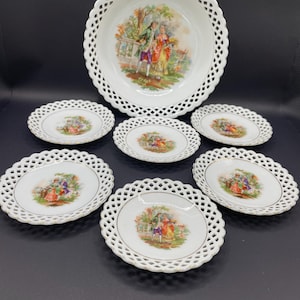 May include: A white porcelain serving set featuring a large bowl and six smaller plates. Each piece has a delicate, openwork lace design around the edges and a central image of a couple in period clothing. The artwork is in shades of green, yellow, red, and purple.