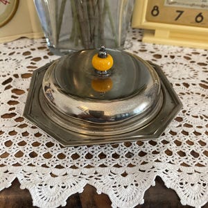 Peut inclure: Un beurrier argenté avec un couvercle et un bouton jaune vif. Le plat a une base octogonale et un couvercle bombé. Le bouton est de couleur jaune vif. Le plat est posé sur un napperon en dentelle blanche.