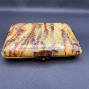 May include: A rectangular, vintage, tortoiseshell-patterned compact case. The case features a warm color palette of yellow, brown, and amber hues. It has a clasp closure and a hinged design, suggesting it opens to reveal a mirror and compartments.