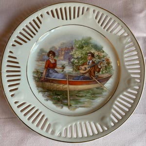 May include: A decorative porcelain plate with a romantic scene of a couple in a rowboat, with a man playing a lute. The plate has a white, pierced rim with a gold trim. The image depicts a serene waterside setting.