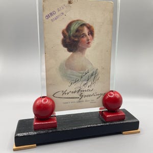 May include: A vintage postcard featuring a woman with a green ribbon in her hair. The postcard is framed in a clear acrylic stand with red wooden accents. The postcard reads "Felice Natale 1915 Christmas Greetings There's such a charm about you."