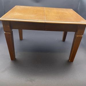 Puede incluir: Una pequeña mesa rectangular de madera con un acabado marrón cálido. La mesa tiene cuatro patas robustas y una tapa de dos piezas. La mesa mide aproximadamente 61 cm de largo, 46 cm de ancho y 51 cm de alto.