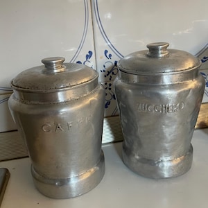 Pode incluir: Dois potes de metal vintage com tampas. Um pote está rotulado "CAFFE" e o outro "ZUCCHERO". Os potes são prateados e parecem ser feitos de alumínio. Os potes são provavelmente para guardar café e açúcar.