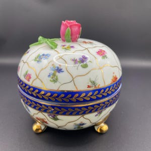 Op de afbeelding: Een ronde, witte porseleinen sieradendoos met een roze roos erop. De doos heeft een blauwe band met bladgoud details en is versierd met kleurrijke bloempatronen en gouden accenten. De doos staat op drie gouden voetjes.