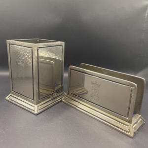 Op de afbeelding: Twee zilverkleurige bureau-accessoires: een vierkante pennenhouder en een briefhouder. Beide items hebben een rechthoekig ontwerp met een decoratief embleem. De pennenhouder heeft een doosvorm, terwijl de briefhouder een schuin ontwerp heeft.