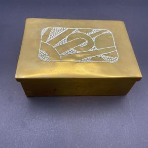 Puede incluir: Una caja rectangular de latón con tapa. La tapa presenta un diseño abstracto blanco con elementos punteados y lineales. La caja tiene un diseño simple y limpio y un color metálico cálido.
