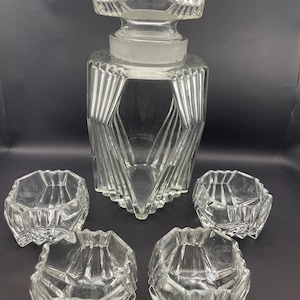 Könnte beinhalten: Eine klare Glasparfümflasche mit geometrischem Design, mit einem Stopfen und vier kleinen passenden Schalen. Die Flasche hat eine facettierte Form mit vertikalen Linien, und die Schalen haben ein ähnliches Design. Das Set wird auf einer dunklen Oberfläche präsentiert.