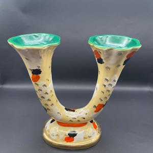 Può includere: Un vaso vintage a forma di U con due beccucci. Il vaso presenta un corpo color crema con accenti neri e arancioni, una base dorata e bordi verdi. Questo pezzo decorativo è probabilmente in ceramica e adatto per composizioni floreali o esposizioni.