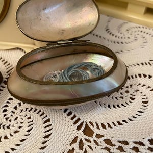 Op de afbeelding: Een open, ovaalvormige sieradendoos met een parelmoer schelpontwerp. De doos heeft een scharnierend deksel en een messingkleurige metalen rand. Binnenin zit een blauw koord en kleine zilveren voorwerpen. De doos staat op een wit gehaakt kleedje.