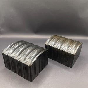 May include: Two dark-coloured, rectangular blocks with a ridged top surface. The blocks have a textured, vertical-lined side. The tops have a series of parallel, curved ridges. The blocks are set against a neutral background.