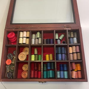 Op de afbeelding: Een houten naaidoos met een glazen deksel, gevuld met spoelen garen in verschillende kleuren, knopen en naaigerei. De doos is verdeeld in compartimenten, met rode stof die de bodem bekleedt. Er zijn verschillende kleuren garen zichtbaar.