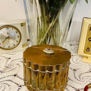 Peut inclure: Un récipient en laiton vintage avec un bord festonné et un bouton décoratif sur le couvercle. Le récipient a une forme cylindrique avec des détails verticaux nervurés. Le laiton a une patine chaude et vieillie. Le récipient repose sur une nappe en dentelle blanche.