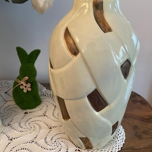 Op de afbeelding: Een crèmekleurige keramische vaas met een geweven ontwerp en bruine accenten. De vaas heeft een glanzende afwerking en een brede, afgeronde vorm. Een groen konijnbeeldje en witte bloemen staan op de achtergrond.
