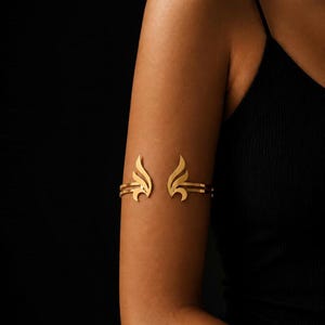 May include: A gold-toned metal arm cuff featuring a symmetrical design of two stylized wing or flame motifs. The open-ended band consists of two parallel thin metal strips that curve outward to form the decorative wing shapes on the upper arm.