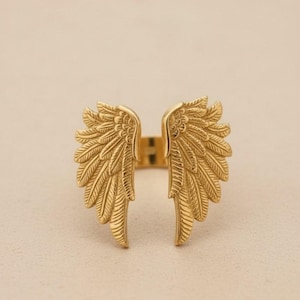 May include: A gold-colored ring featuring an open design with detailed wing motifs. The wings are spread out, with intricate feather detailing. The ring is set against a neutral background, highlighting the jewelry's design.