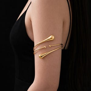 Pode incluir: Punho de braço dourado com um design minimalista. O punho apresenta dois detalhes em forma de gota e duas pequenas esferas. A joia é usada na parte superior do braço contra um fundo preto.