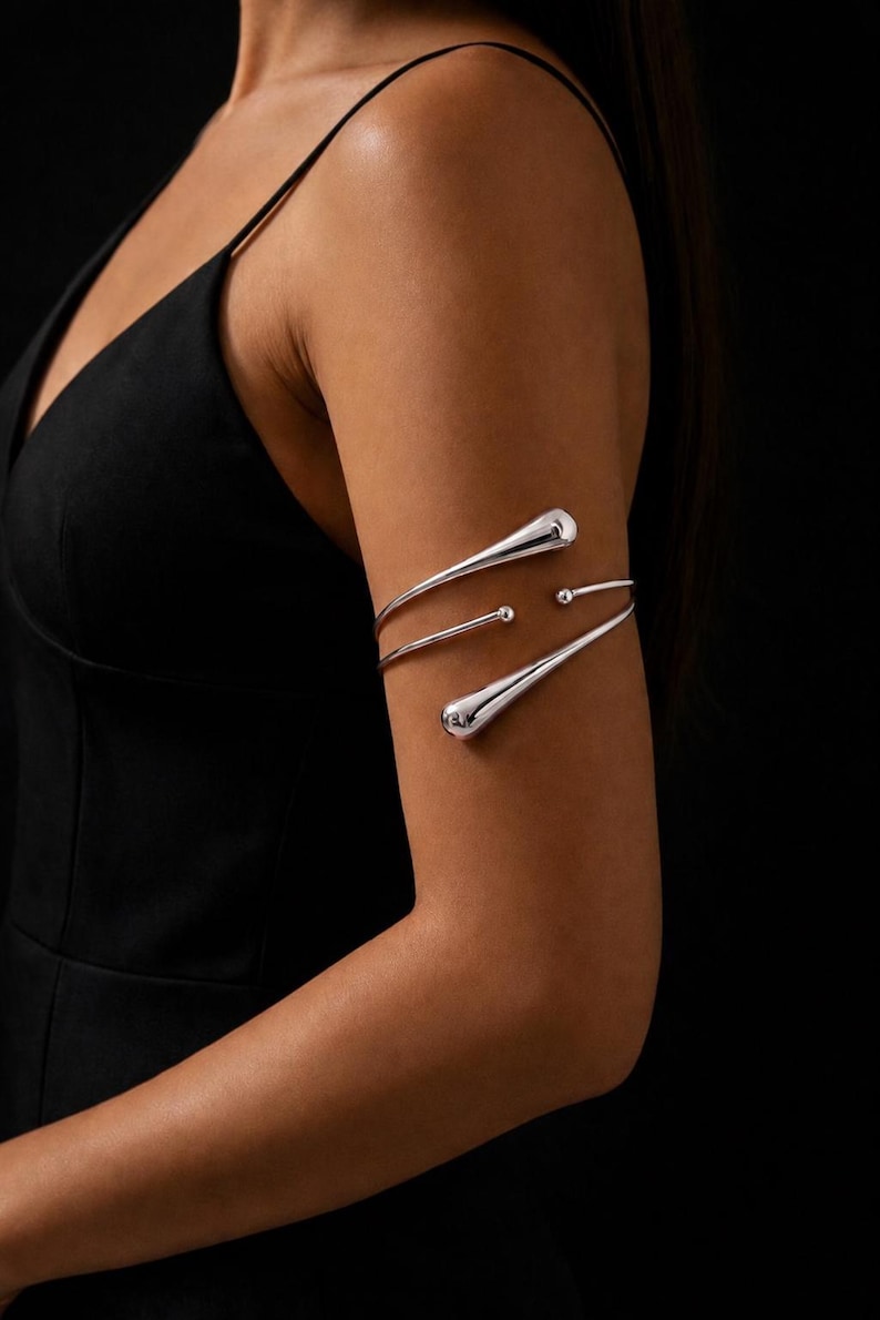 May include: Silver arm cuff with a minimalist design. The cuff features three curved, polished metal bands with teardrop-shaped ends and small spherical accents. The jewelry is worn on the upper arm against a black background.