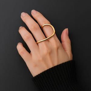 以下が含まれることがあります： ユニークな曲線デザインのゴールドカラーのリング。 リングは2本の指に巻き付き、モダンでミニマルな外観を作り出します。 ジュエリーは、暗い背景に対して手に表示されます。
