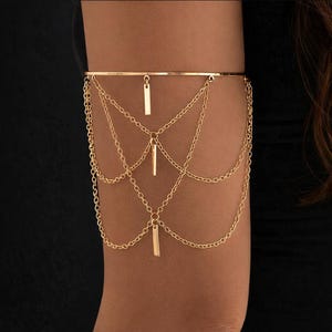 Könnte beinhalten: Goldene Armspange mit horizontalem Stab und drei vertikalen Stabanhängern, verbunden durch zarte Ketten. Der Armschmuck wird an einem Arm vor dunklem Hintergrund getragen. Das Design ist elegant.