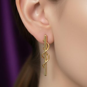 Puede incluir: Pendientes dorados con un diseño de barra vertical y un delicado acento en espiral. Los pendientes se muestran puestos en una oreja, con un fondo morado. Los pendientes tienen un acabado pulido y un estilo minimalista.