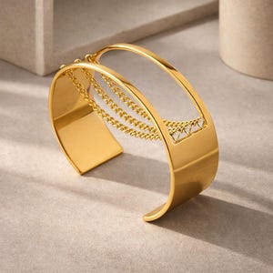 May include: A gold-colored cuff bracelet with a rectangular base and a curved top. Delicate gold chains hang from the top, connecting to the base. The bracelet has a modern, open design, and is displayed on a neutral surface.