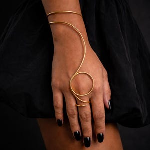 Puede incluir: Un conjunto de pulsera y anillo de mano de color dorado. La pulsera se envuelve alrededor de la muñeca y se conecta a un anillo a través de un diseño curvo. El anillo es una banda simple. La joya se usa en una mano con esmalte de uñas negro.