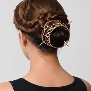 May include: A gold-coloured hair accessory is worn in a bun. The hair accessory has a circular design with intricate swirls and is secured with two long, thin pins. The person is wearing a black vest top.
