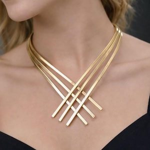 May include: A gold-coloured necklace with a geometric design. The necklace features multiple flat bars that intersect in a layered pattern. The jewellery is worn against a dark background.