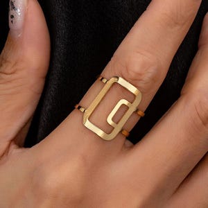 Op de afbeelding: Een goudkleurige ring met een rechthoekig ontwerp. De ring heeft een grote, open rechthoek met een kleinere, verbonden rechthoek aan de binnenkant. De ring wordt om een vinger gedragen en benadrukt de moderne, geometrische stijl. De ring is een sieraad.