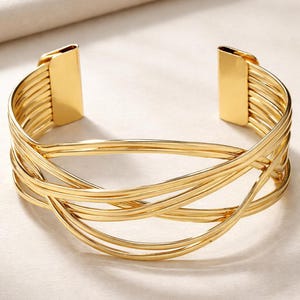Pode incluir: Uma pulseira de punho dourada com um design aberto. A pulseira apresenta várias faixas paralelas que se curvam e se cruzam, criando um padrão entrelaçado. As extremidades da pulseira têm fechos retangulares sólidos.