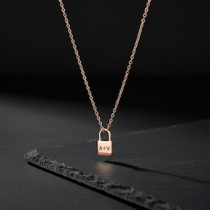 Op de afbeelding: Een roségouden hangslot-hangerketting met een fijne ketting. De hanger heeft de gegraveerde initialen "A+V". De ketting wordt getoond tegen een donkere achtergrond, wat het elegante ontwerp benadrukt.