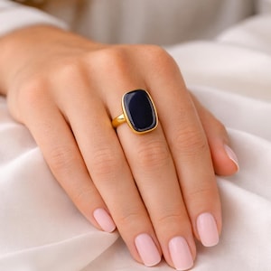 May include: A gold ring with a rectangular, dark blue stone. The ring is set on a hand with pink nail polish, resting on a white satin fabric. The ring's design is simple and elegant.