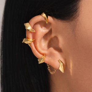 Könnte beinhalten: Goldfarbenes Ohrklemmen-Set mit mehreren blattförmigen und spitzen Designs. Das Set enthält eine Kette, die zwei Teile verbindet und einen geschichteten Effekt erzeugt. Der Schmuck wird am Ohr getragen und zeigt einen modernen und kantigen Stil.