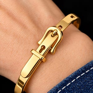 May include: A polished gold-tone metal bangle bracelet designed to resemble a belt, featuring a functional-style buckle closure, a central pin, and a series of small holes along the band, creating a sleek and structured appearance.