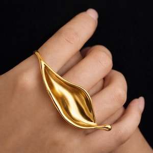 May include: A gold-colored leaf-shaped ring. The ring features a smooth, polished surface and a delicate, elongated design. The ring is worn on a finger, with the leaf shape extending across the top of the hand. The ring's design is simple yet elegant.