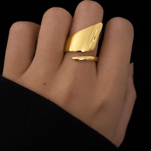 Op de afbeelding: Een goudkleurige ring met een abstract ontwerp. De ring heeft een groot, hoekig bovendeel en een dunnere band. De ring wordt getoond tegen een donkere achtergrond. Het ontwerp van de ring is modern en opvallend.