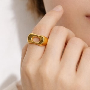 May include: A gold ring with a rectangular design and a central void. The ring is on a finger, showcasing a modern, minimalist aesthetic. The polished surface of the ring reflects light, creating a bright, shiny appearance.