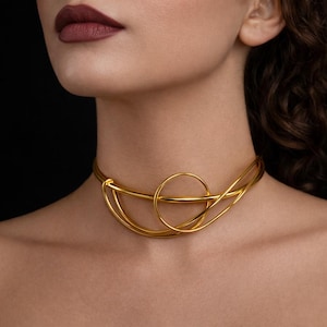 Könnte beinhalten: Ein goldfarbenes Choker-Halsband mit einem abstrakten Design. Die Halskette hat ein kreisförmiges Element in der Mitte, mit geschwungenen Linien, die sich um den Hals erstrecken. Der Schmuck wird vor einem dunklen Hintergrund präsentiert.