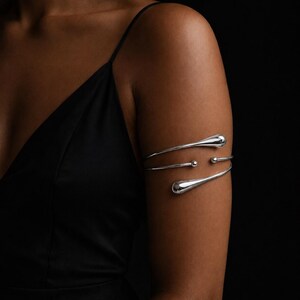 May include: Silver arm cuff with a teardrop design. The cuff wraps around the upper arm with three curved bands, featuring teardrop-shaped ends and small spherical accents. The jewelry is displayed against a dark background.