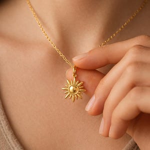 Puede incluir: Un collar con colgante de sol dorado. El colgante de sol tiene un centro redondo con rayos que se extienden hacia afuera. La cadena del collar también es dorada. El collar está siendo sostenido por una persona.